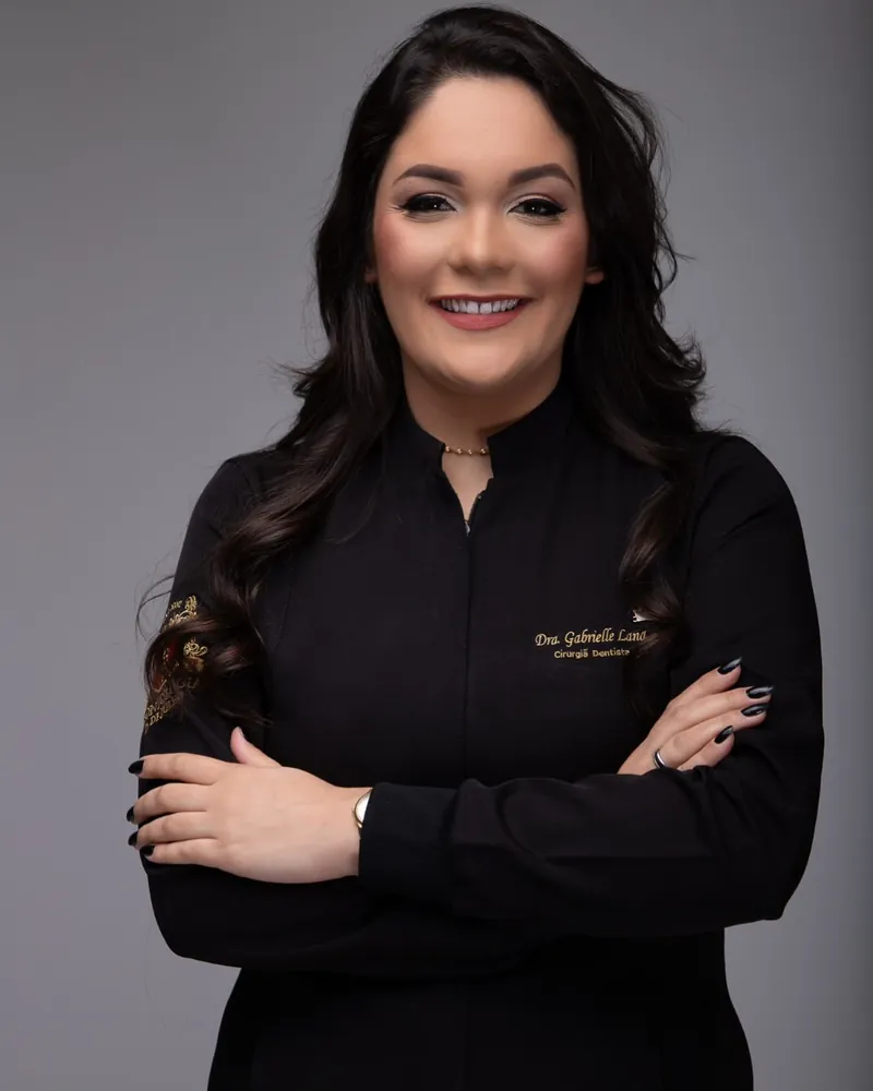 Dra. Gabrielle Lana, de jaleco preto, sorrindo de braços cruzados