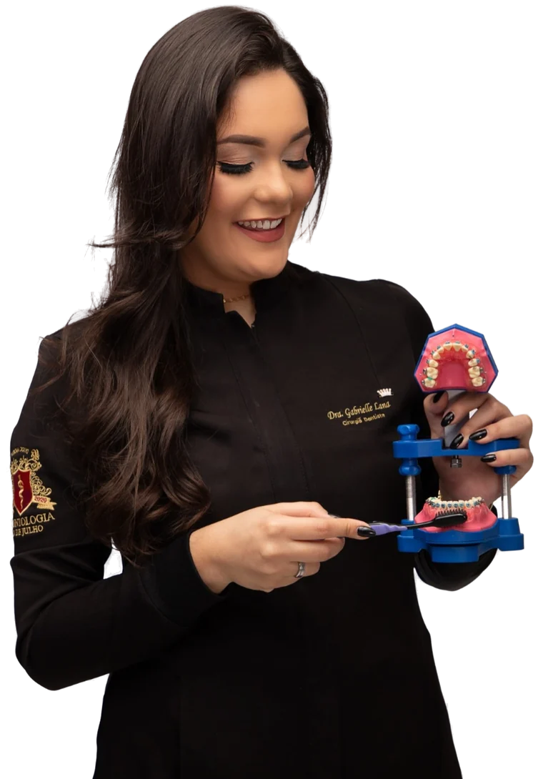 Dra. Gabrielle Lana, de jaleco preto, sorrindo enquanto mostra a escovação em um modelo de arcada